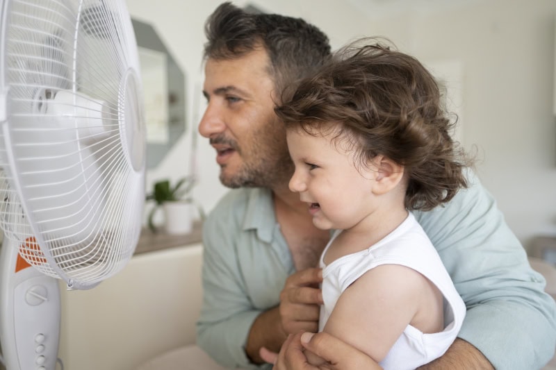 Father and child is front of electric fan on hot summer day.