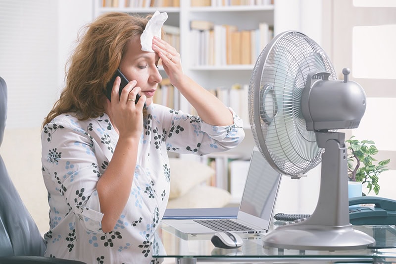 When Do I Replace My Air Conditioner? A woman sitting in front of a fan while wiping the sweat from her forehead.