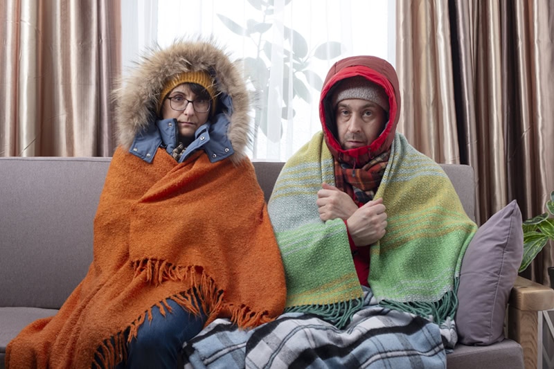 A man and a woman sitting side by side on a sofa, looking cold and uncomfortably shivering indoors.