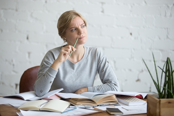 Woman sitting at a table, with books open. She has a pensive look on her face.