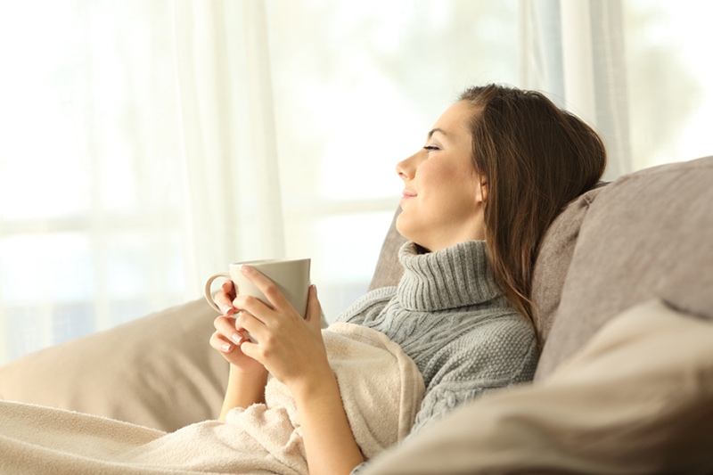 A woman relaxing on her couch with a blanket & a cup of coffee.