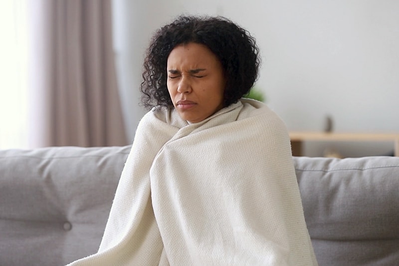 Woman wrapped in a blanket shivering on her couch.