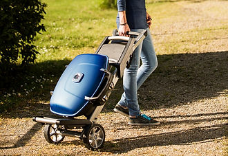 Travel barbecue being wheeled outside on path by a gentleman wearing blue jeans and a navy shirt.